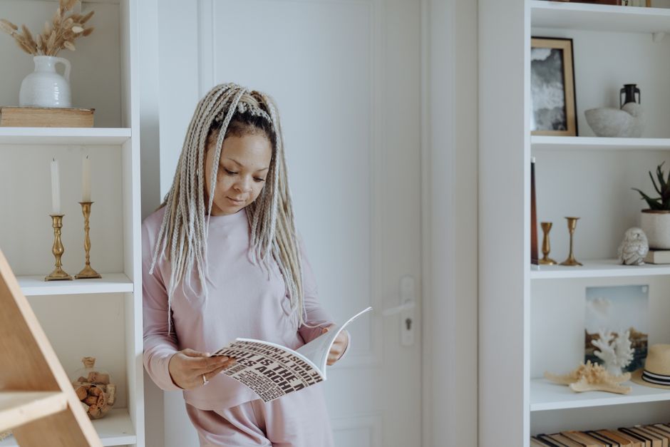 Black woman with long braids reading in stylish home interior, against white background.